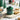 Green ceramic jar with decorative lid on a wooden board with gingerbread cookies, Christmas trees, and ornaments in the background.