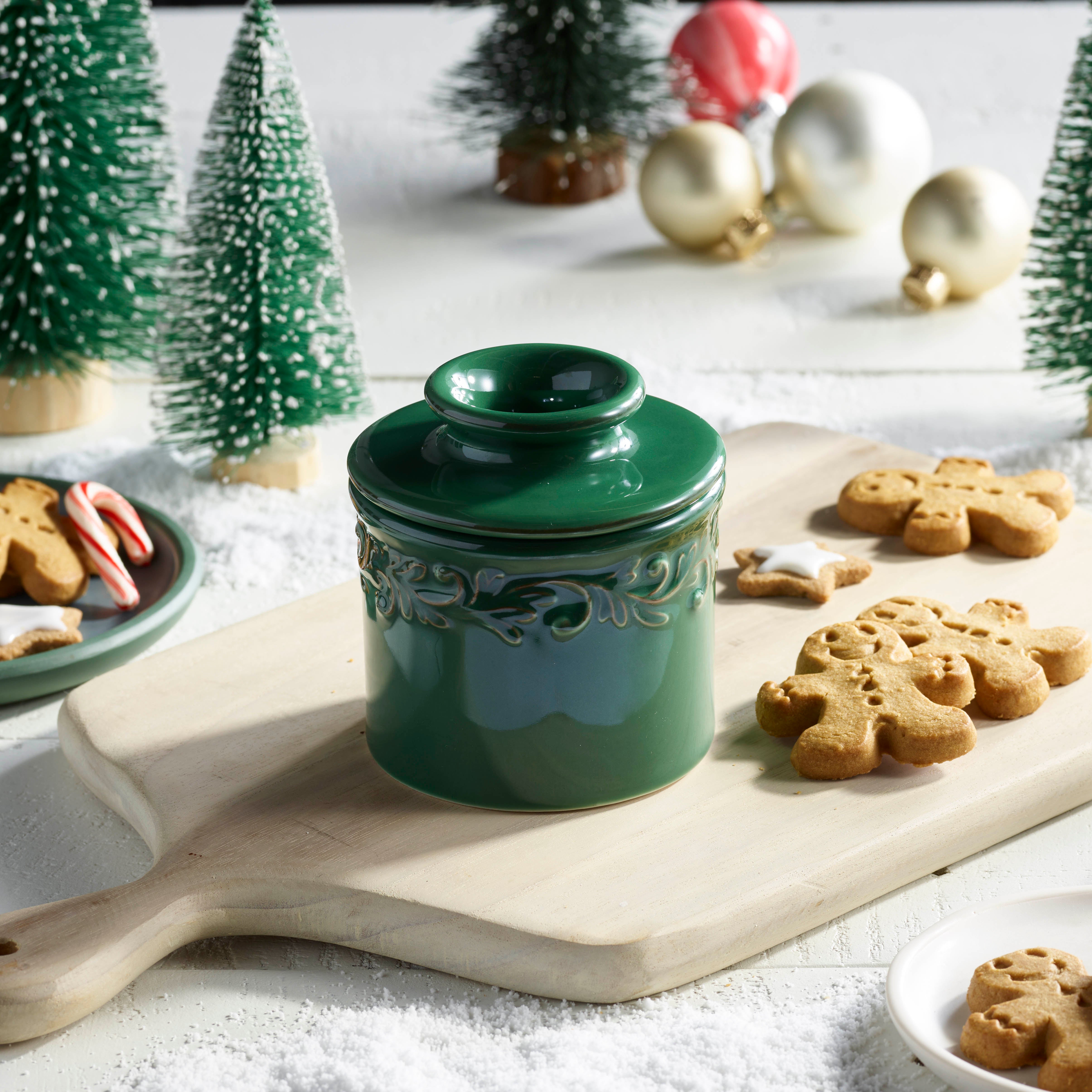 Green ceramic jar with decorative lid on a wooden board with gingerbread cookies, Christmas trees, and ornaments in the background.